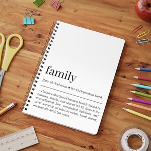 May include: A white spiral-bound notebook with the word "family" and its definition printed on the cover. The notebook is on a wooden desk, surrounded by stationery items such as pencils, scissors, and paper clips.