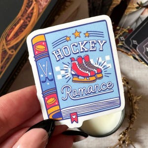 May include: A sticker with a blue background featuring the words "HOCKEY Romance" and an illustration of red ice skates. The sticker has a book-like design with hockey sticks and pucks on the spine.