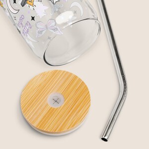 May include: A clear glass tumbler with a bamboo lid and a silver metal straw. The glass is decorated with celestial and butterfly designs. The words "little osha" are printed on the glass.