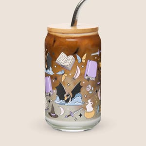 Op de afbeelding: Een doorzichtig, blikvormig glas gevuld met ijskoffie, voorzien van een bamboe deksel en een metalen rietje. Het glas is versierd met kleurrijke illustraties van een draak, open boeken, vlinders, een ketel, een zwaard en een heksenhoed.