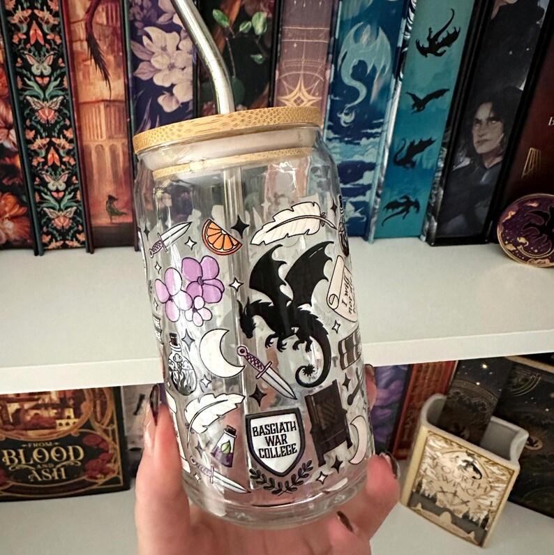 May include: A clear glass tumbler with a bamboo lid and a metal straw. The tumbler is decorated with various black and white illustrations, including a dragon, flowers, and the text "Basgiath War College".