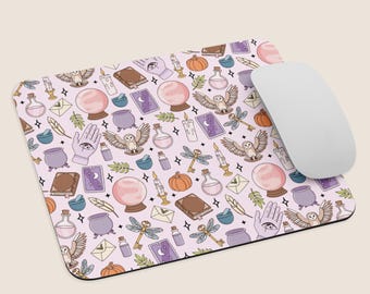 Puede incluir: Alfombrilla de ratón rectangular con fondo rosa claro y un patrón repetido de elementos místicos. El patrón incluye bolas de cristal, búhos, botellas de pociones, velas y otros símbolos ocultos. Un ratón de ordenador blanco descansa en el lado derecho.