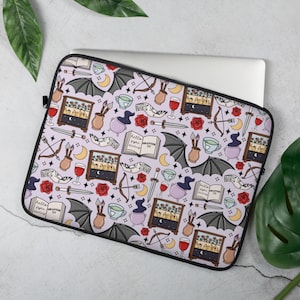 May include: A laptop sleeve featuring a repeating pattern of fantasy-themed illustrations. The design includes bats, moons, bows and arrows, potion bottles, roses, and open books with the text "Hello figue darling" and "Chapter 33". The background is a pale lilac colour.