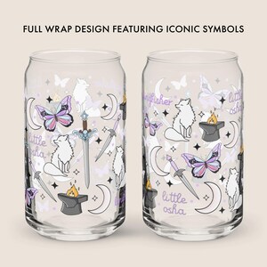 May include: Two clear glass can-shaped cups with a wraparound design featuring iconic symbols. The design includes butterflies, crescent moons, swords, foxes, and text that reads "little osha" and "kingfisher". The cups are decorated with purple, white, and gold accents.