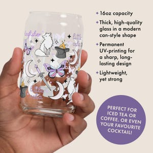 May include: A clear 16oz can-shaped glass with a whimsical design featuring white wolves, butterflies, moons, and a sword. The glass has the words "little osha" and "ingfisher" printed on it. The glass is held in a hand.