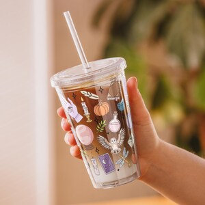 May include: A clear plastic tumbler with a lid and straw, filled with a layered drink. The tumbler is decorated with colorful illustrations of a hand, candle, pumpkin, owl, and other mystical symbols. The tumbler is held in a person's hand.