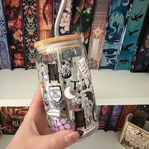 May include: A clear glass tumbler with a bamboo lid and a stainless steel straw. The tumbler is decorated with fantasy-themed illustrations, including swords, potions, and the text "BASGIATH WAR COLLEGE" and "I will not die today".