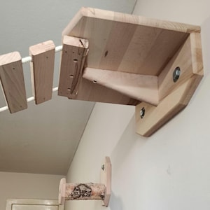 Op de afbeelding: Een houten kattenklimstructuur met een plank en een touwbrug. De plank is met metalen hardware aan de muur bevestigd. De brug is gemaakt van houten blokken en een wit touw. De structuur is gemaakt van lichtgekleurd hout.