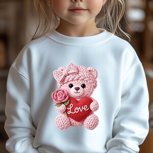 May include: White sweatshirt featuring a pink knitted teddy bear holding a red heart with the word "Love" and a pink rose. The teddy bear has a bow on its head. The sweatshirt is for children.