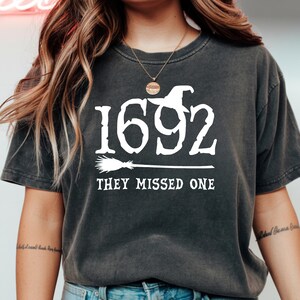 May include: Dark gray t-shirt with the white text "1692 THEY MISSED ONE". The design includes a witch hat and broom, evoking a Halloween or witch theme. The shirt has a vintage, worn look.