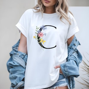 May include: White t-shirt featuring a large black letter "C" adorned with a floral arrangement in pink, yellow, and purple. The shirt has rolled-up sleeves and is paired with a denim jacket.