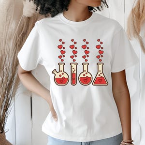 May include: White t-shirt featuring a graphic of four laboratory flasks with red liquid and heart-shaped bubbles rising from each. The flasks have a beige outline and a heart inside. A fun, science-themed design.