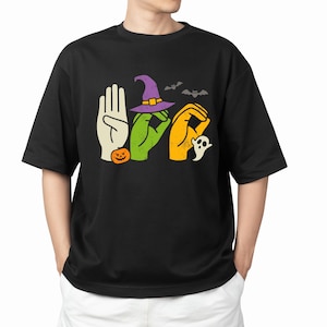 Könnte beinhalten: Schwarzes T-Shirt mit einem Halloween-Motiv. Das Design buchstabiert "BOO" mit Handgesten, einem Hexenhut, einem Kürbis, einem Geist und Fledermäusen. Das T-Shirt ist ein lässiger Stil mit kurzen Ärmeln.