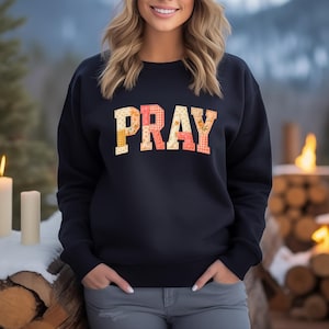 Könnte beinhalten: Ein marineblaues Sweatshirt mit dem Wort "PRAY" in einem Patchwork-Design. Die Buchstaben sind mit Blumen- und Vichy-Mustern in Gelb-, Rosa- und Orangetönen gefüllt. Das Sweatshirt hat einen Rundhalsausschnitt.