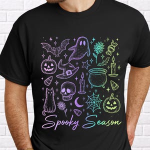 May include: A black short-sleeve t-shirt featuring a colorful graphic print of Halloween icons including a ghost, bat, cat, skull, pumpkin, cauldron, and spiderweb. The text "Spooky Season" is printed in a gradient script font at the bottom.
