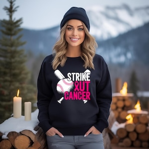 Könnte beinhalten: Ein marineblaues Sweatshirt mit dem Schriftzug "Strike Out Cancer" in Weiß und Rosa, mit einer Baseball- und Schlägergrafik. Das Sweatshirt wird von einer Person getragen, die eine marineblaue Mütze und eine graue Hose trägt.