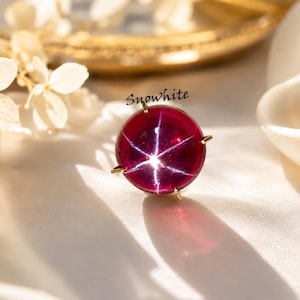 May include: A round, deep red star ruby gemstone set in a gold prong setting. The stone displays a distinct six-rayed star effect across its surface. The word "Snowhite" is printed in black script text above the gemstone.