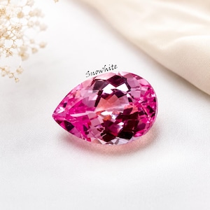 Puede incluir: Una gema rosa facetada en forma de pera con la palabra "Snowwhite" en escritura cursiva. La gema está sobre un fondo blanco y crema, con una ramita de flores blancas en la esquina superior izquierda.