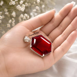 May include: A hexagonal pendant featuring a deep red faceted gemstone set in a silver-toned metal frame with a decorative filigree bail. The word Snowhite is printed in black script lettering near the top edge of the red gemstone surface.