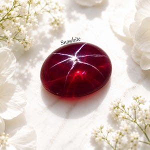 May include: A polished, oval-shaped, deep red gemstone with a star-like pattern of white lines radiating from the center. The word "Snowwhite" is written above the gem. White flowers and greenery surround the gem on a white surface.