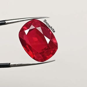 May include: A vibrant red gemstone, held by tweezers, with the word "Snowwhite" in white text. The gem has a cushion cut with multiple facets, reflecting light. The background is a plain white.