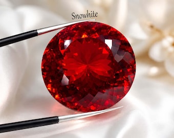 May include: A large, faceted, round, red gemstone, held by tweezers. The gem has many facets that reflect light, creating a brilliant sparkle. The word "Snowwhite" is written above the gem.