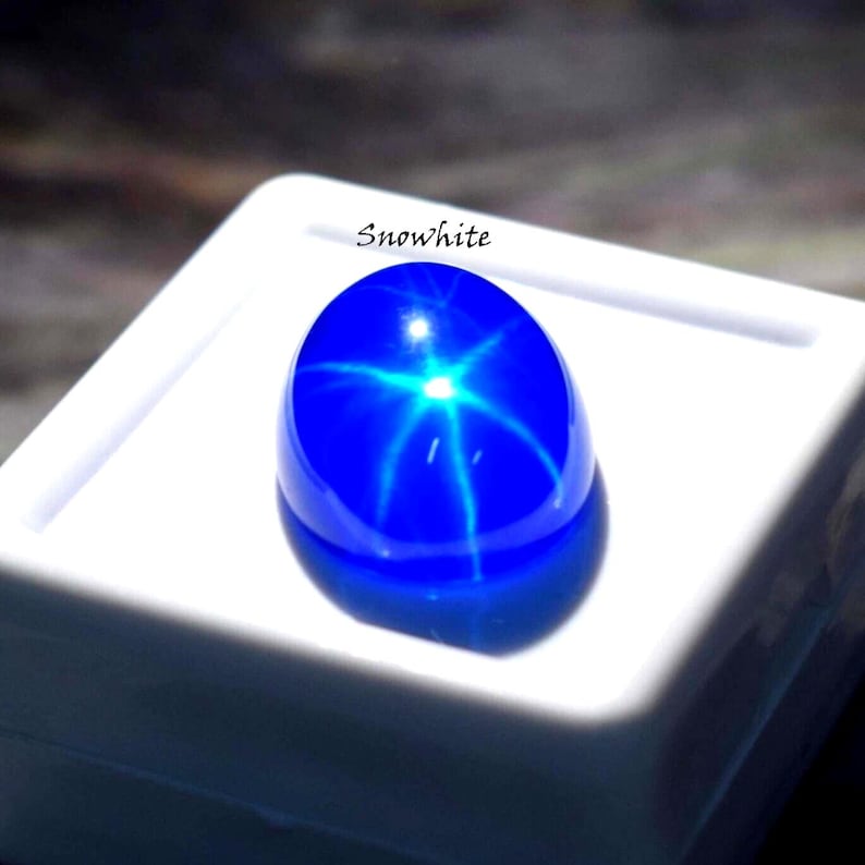 Puede incluir: Una gema cabuj&oacute;n ovalada de color azul vibrante que presenta un efecto de estrella de seis rayos, conocido como asterismo, centrado en su superficie lisa y pulida. La piedra se muestra sobre una base cuadrada blanca con el texto Snowhite.