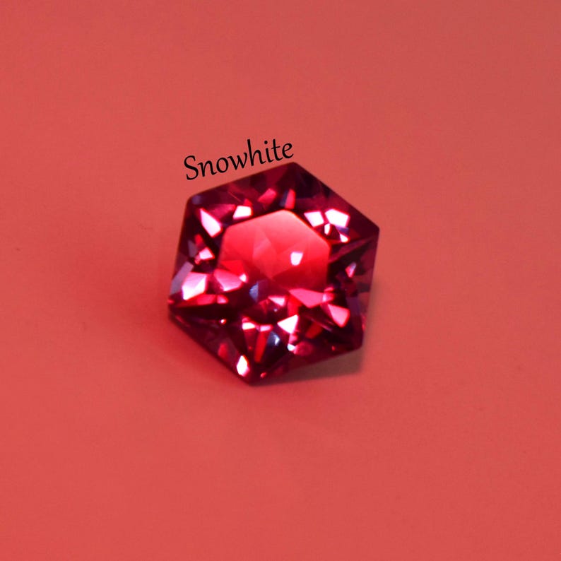 May include: A faceted hexagonal gemstone in a deep magenta-red color with a brilliant cut that reflects light. The word Snowwhite is printed in black text above the gemstone. The object is displayed against a solid, flat coral-colored background.