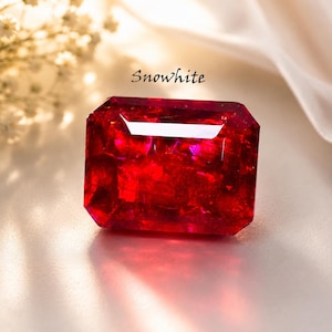 May include: A vibrant, rectangular, red gemstone with the word "Snowwhite" in cursive script. The gem has a reflective surface and is set against a soft, neutral background. The gem is a deep shade of red.