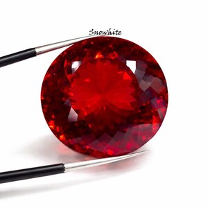May include: A large, faceted, round, deep red gemstone held by tweezers. The gem has many facets that reflect light, creating a brilliant appearance. The word "Snowwhite" is written above the gem.