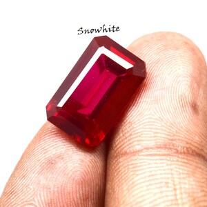 Può includere: Una gemma di rubino rettangolare e sfaccettata, di colore rosso intenso. La gemma è tenuta tra le dita, con la parola "Snowwhite" scritta sopra. Il taglio della pietra permette alla luce di riflettersi, creando un aspetto brillante.