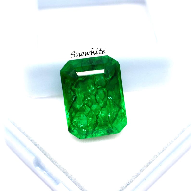 May include: A vibrant, emerald-cut green gemstone with the word "Snowwhite" printed on the top. The gemstone is a vivid green color and has internal inclusions. The gem is set against a white background.