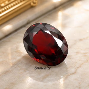 Può includere: Una gemma rosso scuro dal taglio ovale con una superficie sfaccettata che riflette la luce. La pietra è posizionata su una superficie chiara con venature visibili. La parola Snowwhite è stampata in piccolo testo nero sotto la gemma.