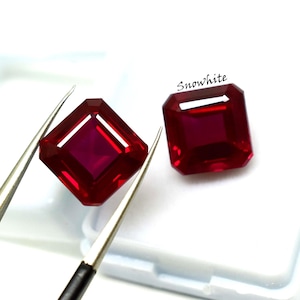 May include: Two radiant-cut, deep red gemstones. One is held by tweezers, showcasing its facets. The other rests beside it. The gems have a vibrant, saturated color and a polished finish. The word "Snowwhite" is written in cursive.