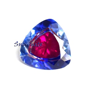 May include: A faceted gemstone with a triangular shape. The gem features a vibrant fuchsia center surrounded by a deep blue outer layer. The word "Snowwhite" is written in cursive on the left side of the gem.
