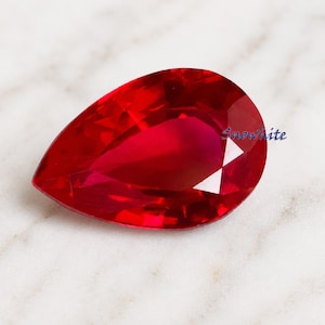 Può includere: Una gemma rossa sfaccettata a forma di pera con un aspetto brillante e trasparente. La pietra mostra profonde tonalità cremisi e nette sfaccettature geometriche che riflettono la luce. La parola Snowwhite è stampata in piccolo carattere blu.