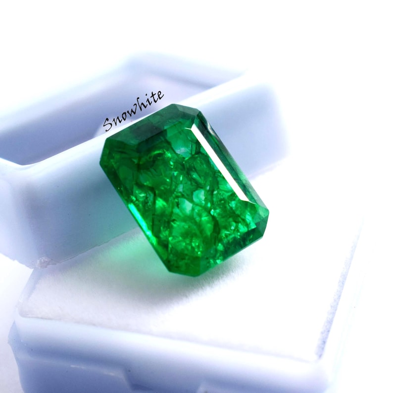 May include: A vibrant, emerald-cut green gemstone with internal inclusions, resting in a white box. The gem is faceted, reflecting light and showcasing its clarity. The word "Snowwhite" is printed on the box.