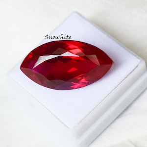 May include: A faceted, marquise-cut ruby gemstone, displayed on a white surface. The gem is a deep, vibrant red with multiple reflective facets. The word "Snowwhite" is printed in black cursive on the upper left.