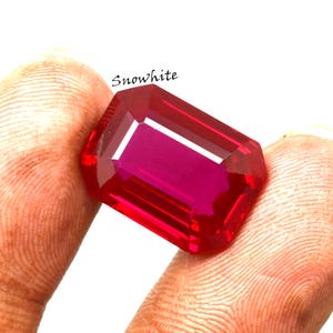 May include: A large, rectangular-cut ruby gemstone with a deep red hue. The gem is held between fingers, showcasing its facets and internal color. The word "Snowwhite" is written in cursive.