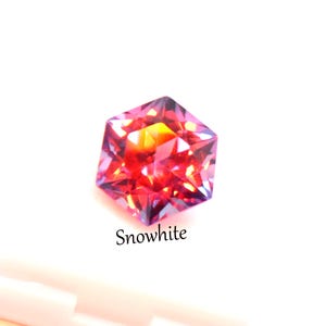 May include: A faceted hexagonal gemstone displaying vibrant shades of red, orange, and purple with internal light reflections. The word Snowwhite is printed in black text below the stone, which is positioned above a white surface.