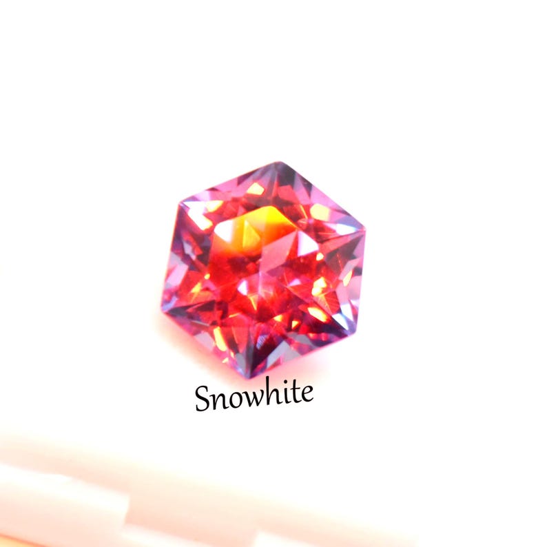 May include: A faceted hexagonal gemstone displaying vibrant shades of red, orange, and purple with internal light reflections. The word Snowwhite is printed in black text below the stone, which is positioned above a white surface.