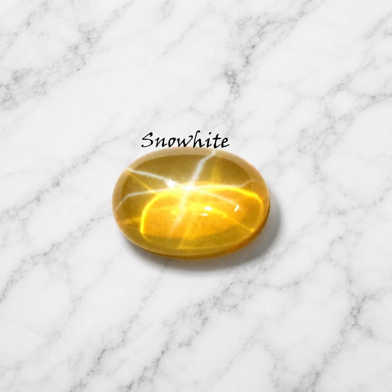 K&ouml;nnte beinhalten: Ein ovaler, goldgelber Edelstein mit einem ausgepr&auml;gten sechsstrahligen Sterneffekt, bekannt als Asterismus. Der Stein hat eine glatte, polierte Cabochon-Oberfl&auml;che und ist mit dem Text Snowwhite direkt &uuml;ber dem Edelstein beschriftet.