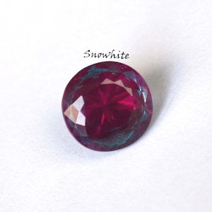 Peut inclure: Une pierre précieuse ronde et facettée, d'un rouge bordeaux profond, avec des reflets de turquoise et de violet. Le mot "Snowwhite" est écrit au-dessus de la gemme en cursive. La gemme est sur fond blanc.