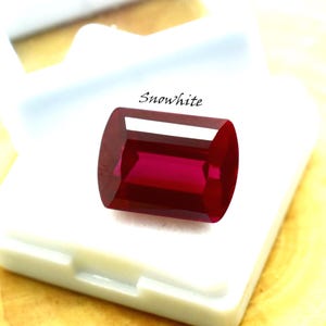 May include: A rectangular, deep red gemstone with a polished surface, set in a white case. The gemstone has a vibrant, saturated colour and is labelled "Snowwhite". The gem is likely a ruby or similar precious stone.