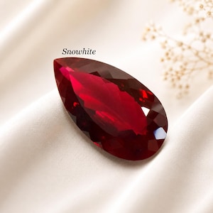 Pode incluir: Uma pedra preciosa facetada em forma de pera de cor vermelha profunda e vibrante. A pedra apresenta um corte brilhante com múltiplas facetas triangulares e em forma de pipa que refletem a luz. A palavra Snowwhite está impressa em preto.