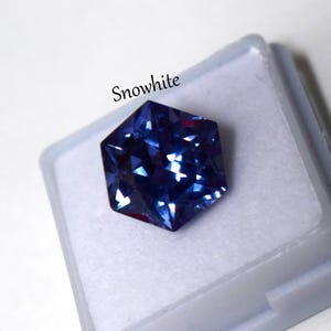 May include: A faceted hexagonal gemstone in a deep blue hue with light-reflecting surfaces, resting inside a small white plastic display case. The word Snowwhite is printed in black text above the gemstone on the white background.