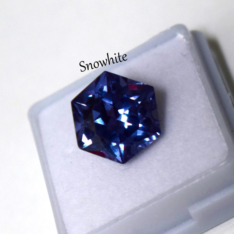 May include: A faceted hexagonal gemstone in a deep blue hue with light-reflecting surfaces, resting inside a small white plastic display case. The word Snowwhite is printed in black text above the gemstone on the white background.
