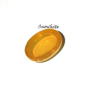 K&ouml;nnte beinhalten: Ein ovaler, durchscheinender bernsteinfarbener Cabochon mit einer glatten, polierten Oberfl&auml;che. Das Wort Snowwhite ist in schwarzer Schreibschrift &uuml;ber dem Objekt abgedruckt. Der Artikel hat einen warmen, goldgelben Farbton und eine gew&ouml;lbte Oberseite.