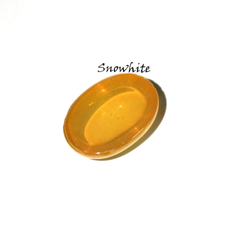 K&ouml;nnte beinhalten: Ein ovaler, durchscheinender bernsteinfarbener Cabochon mit einer glatten, polierten Oberfl&auml;che. Das Wort Snowwhite ist in schwarzer Schreibschrift &uuml;ber dem Objekt abgedruckt. Der Artikel hat einen warmen, goldgelben Farbton und eine gew&ouml;lbte Oberseite.