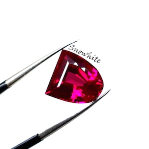 Op de afbeelding: Een gefacetteerde, hartvormige, dieprode edelsteen wordt vastgehouden door een zilveren pincet tegen een witte achtergrond. Het woord "Snowwhite" staat cursief geschreven rechtsboven in de afbeelding.